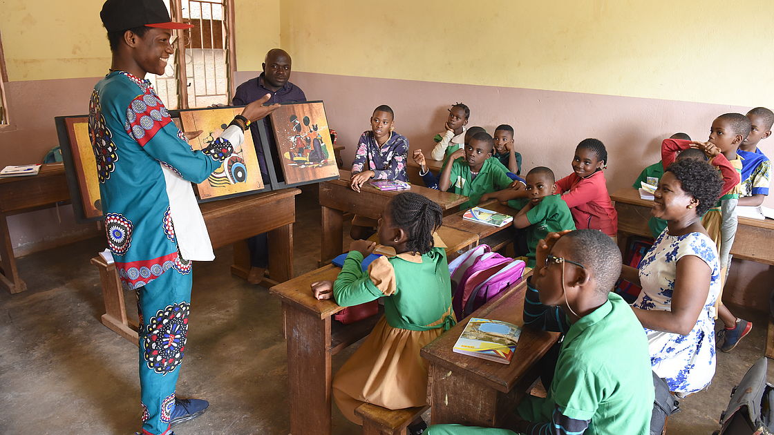 A young man is standing in front of a group of children in a class. He is showing them his artwork. 