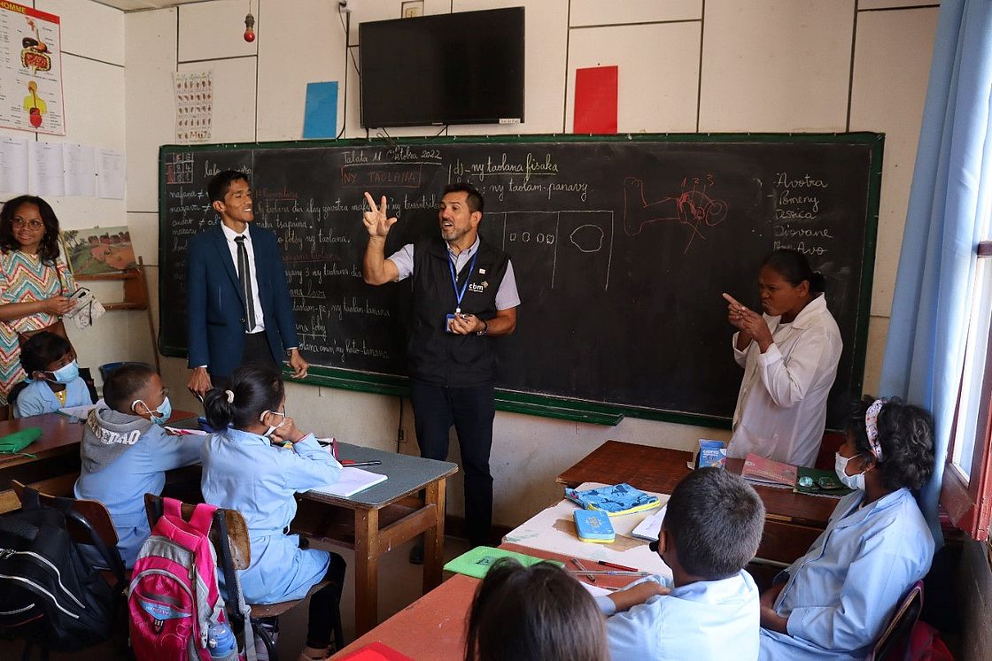 Four people are standing in front of a classroom of students. One man is using sign language to communicate to the students