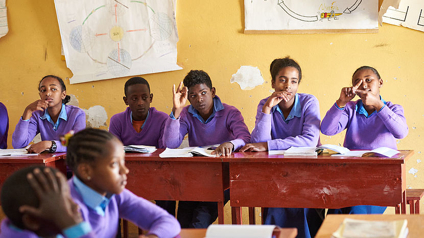 This image shows children in a classroom in Ethiopia, wearing a purple uniform. Some are signing with their hands.