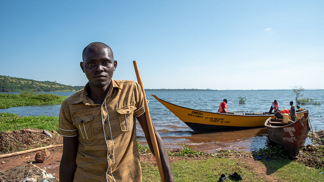 This photo shows a young man standing in front of a boat, he has a long stick in his hand that he uses as a crutch. 