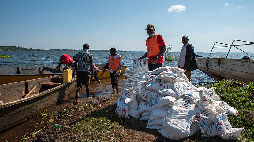 This photo shows a boat full of food material and packets, labelled CBM. Some men and unloading the boat. The Lake Victoria can be seen in the background.