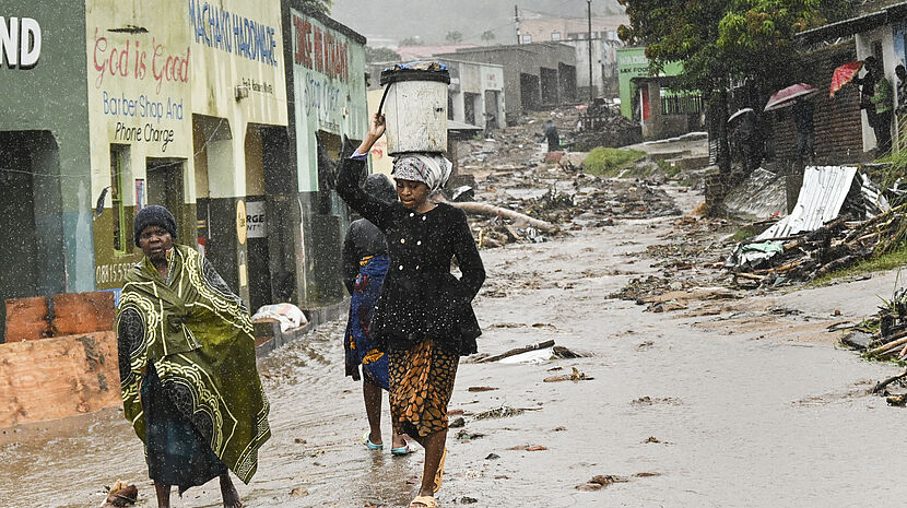 Three people walking under the rain on a flooded road filled with debris