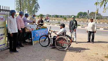 This photo shows a man in a tricycle approaching a cart full of fruits and vegetables at the side of a busy road. There are other men nearby. Everyone is wearing a mask and gloves. 