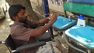 This photo shows a man sitting in a wheelchair, washing his hands at a wash basin with liquid soap.