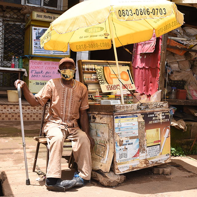 This photo shows a Cameroonian man in local attire sitting next to a mobile kiosk.