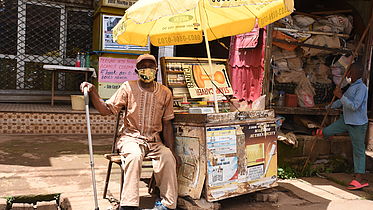 This photo shows a Cameroonian man in local attire sitting next to a mobile kiosk.