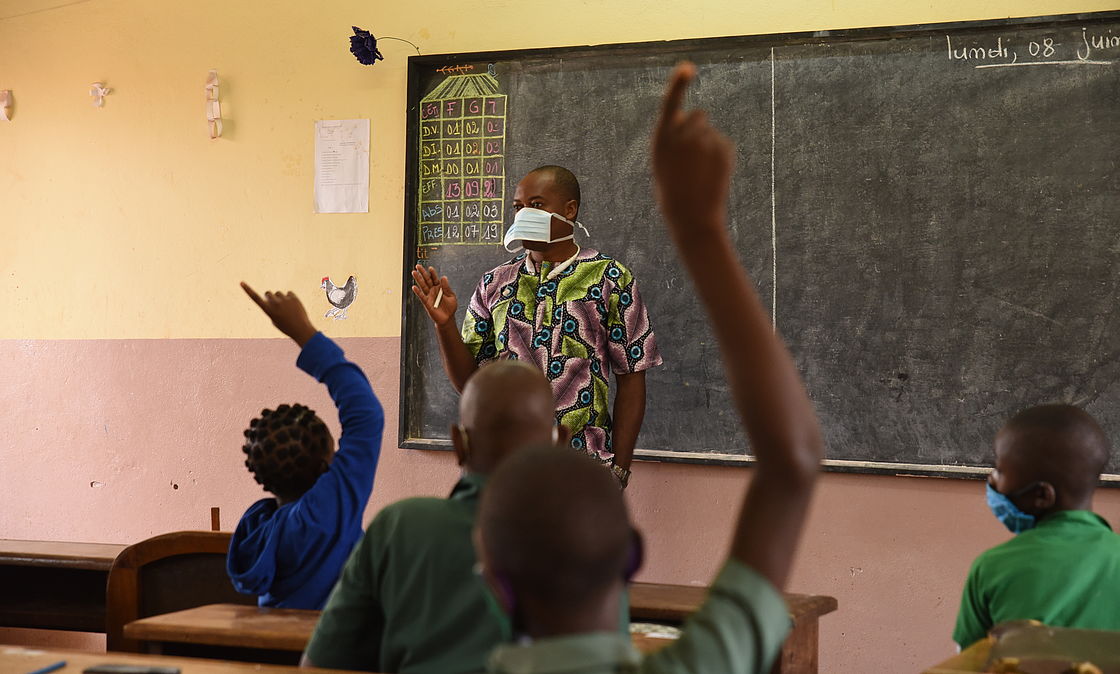 This photo shows a class teacher wearing a mask standing in front of the black board. Several students have their hands raised, to answer the teacher's questions.