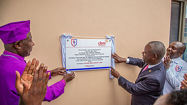 The picture shows a significant moment during the unveiling of a commemorative plaque at the Mengo Eye Complex. Three people are involved in the ceremony: on the left the Archbishop of Uganda, wearing a purple clerical shirt and cassock, on the right the Deputy Speaker of Parliament in a suit and next to him a doctor in a white lab coat. They are outside, next to a light, peach-coloured wall. The Archbishop and the Deputy Speaker of Parliament are unveiling the plaque with the emblems and the text indicating the commissioning of the complex, which was financed by the Christian Blind Mission (CBM) and inaugurated by prominent Ugandan church and government representatives. A small crowd, partially visible, claps and celebrates this milestone in the background.