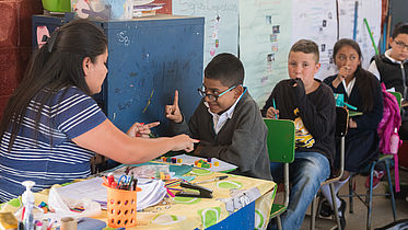 This photo shows a young boy using sign language to communicate with his teacher in a classroom. Other students are also seen sitting at their desks.