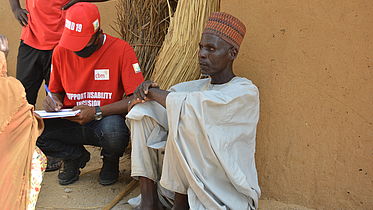 This photo shows an elderly Nigerian man sitting on his haunches, with a staff member next to him, who is carrying a registration board and writing something on it.