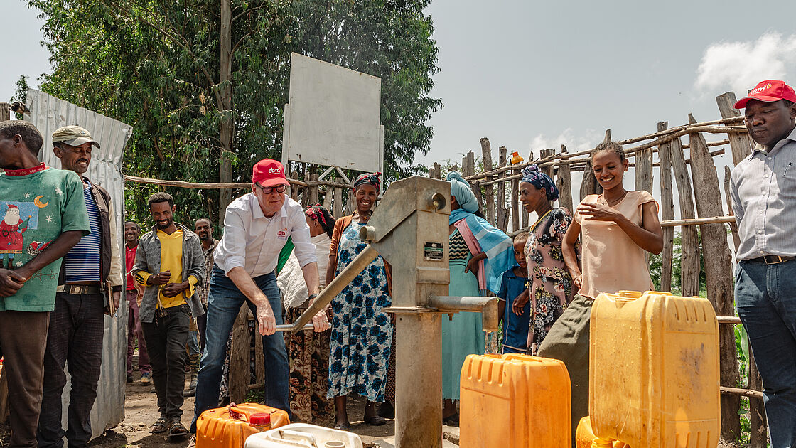 The photo captures a lively moment at a water pump in a rural community, where a diverse group is gathered. A man, distinguishable by his red cap and white shirt, is operating the hand pump, filling yellow jerry cans with water. Surrounding him is a group of residents, including women in traditional attire and a man in a unique Christmas-themed sweater, despite the sunny weather indicating it’s not the winter season. The smiles and attentive faces suggest a positive and interactive atmosphere, possibly a community event or a demonstration of the water pump. The background features simple structures and greenery, typical of a rural landscape.