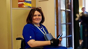 A woman sitting in a wheelchair. She is wearing a blue dress and a smile. 