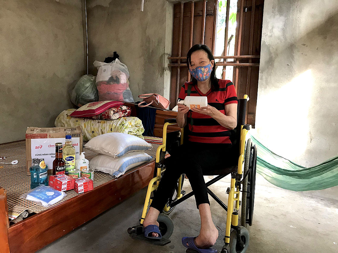 This photo shows a woman in a wheelchair surrounded with some relief material like food and hygiene items. She is wearing a mask. 