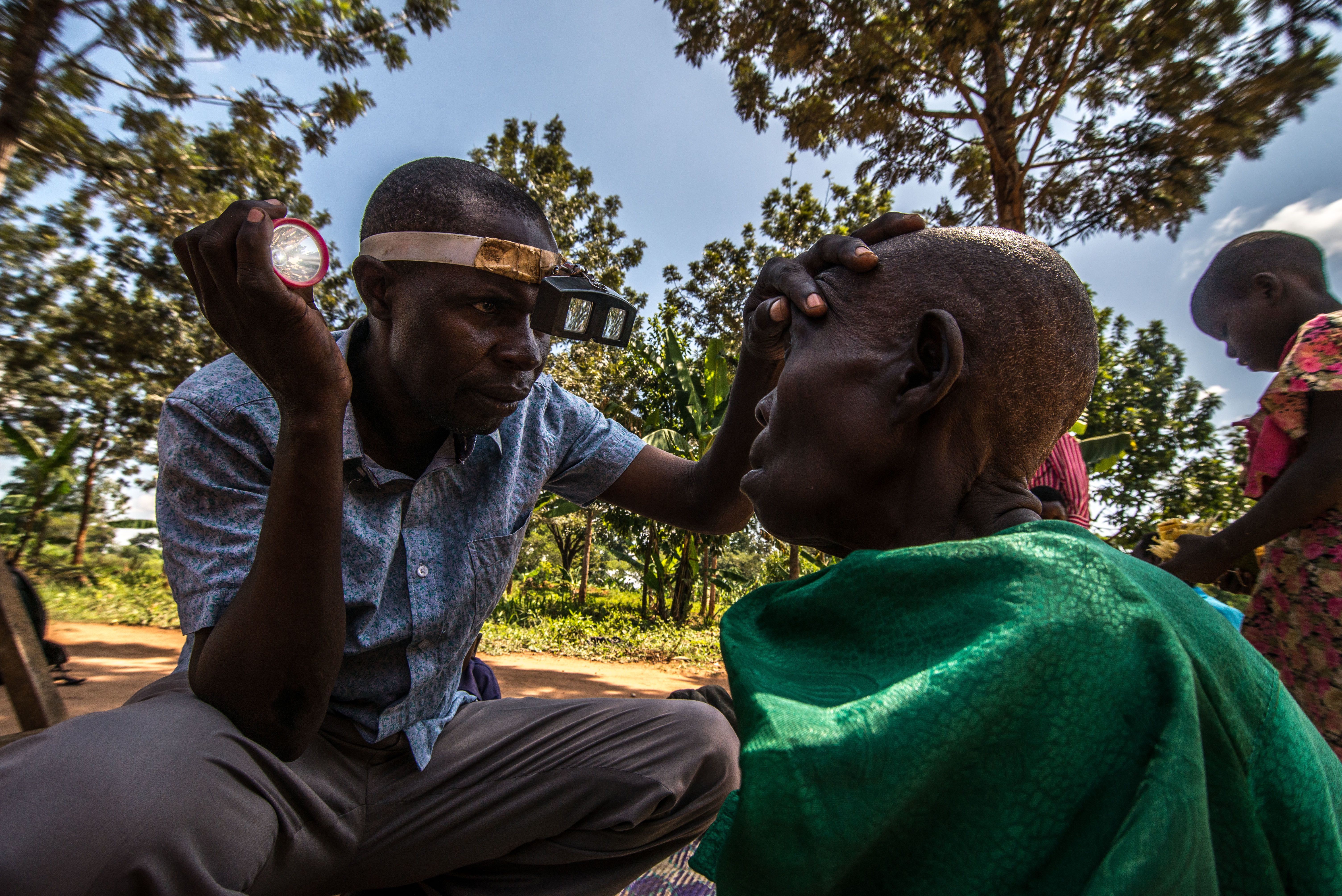 This photo shows a Ugandan man inspecting an old Ugandan woman's eyes in a field. 