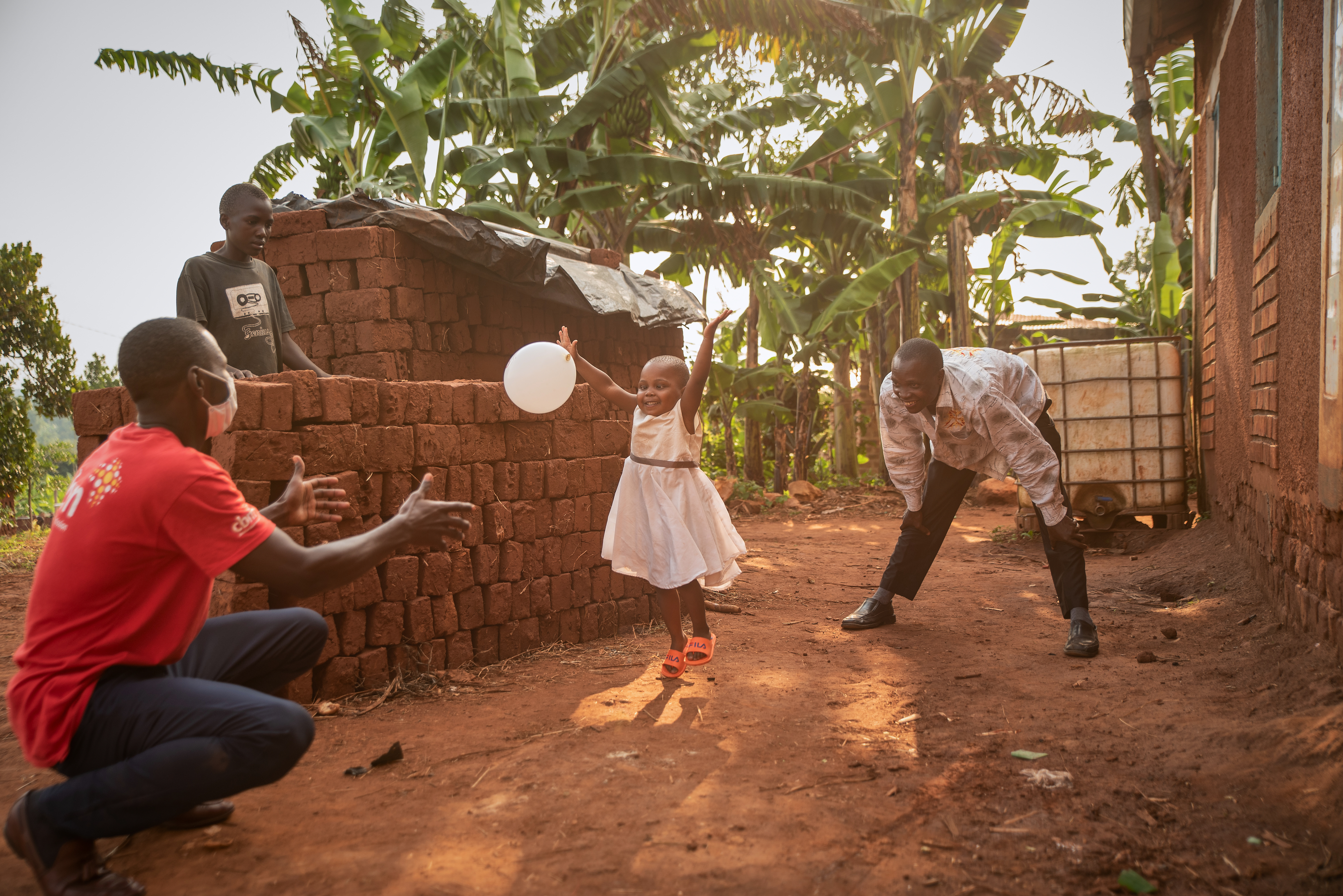 This photo shows a 3 year old girl wearing a white dress playing with a balloon with an African man wearing a CBM Tshirt.